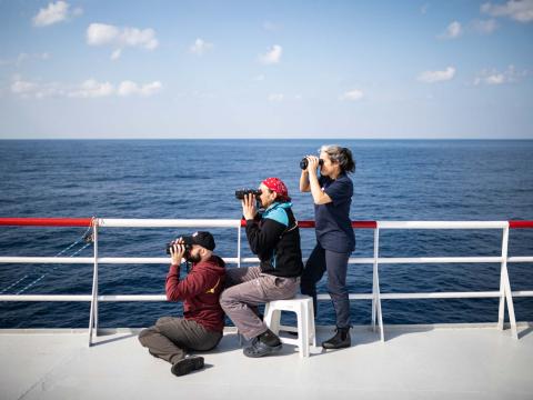 Trois membres de l'équipage observent la mer a l'aide de jumelles depuis la plateforme la plus élevée du bateau. Deux d'entre eux ont été appelés en renfort par un de leur collègue qui vient de repérer une embarcation en détresse.
