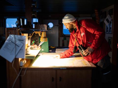 Un membre de l'équipe de recherche et de sauvetage observe une carte maritime sur la passerelle du bateau alors que le jour commence à se lever à l’extérieur.