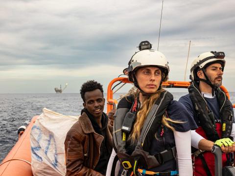 Deux membres de l'équipe de recherche et de sauvetage se tiennent à bord d'un semi-rigide durant un sauvetage. Un homme qui vient d'être secouru est assis à l'arrière du bateau et une plateforme pétrolière peut être aperçue a l'arrière-plan.