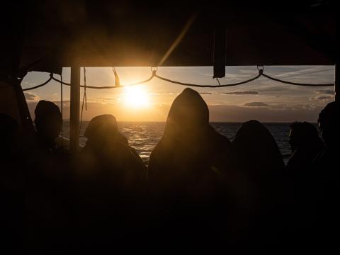 Des rescapés enroulés dans des couvertures regardent le soleil se lever, l'horizon et la mer depuis le pont du bateau.